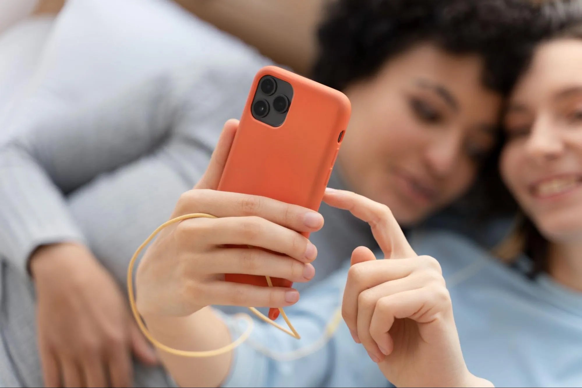 Duas mulheres deitadas juntas segurando um smartphone com capinha laranja vibrante.