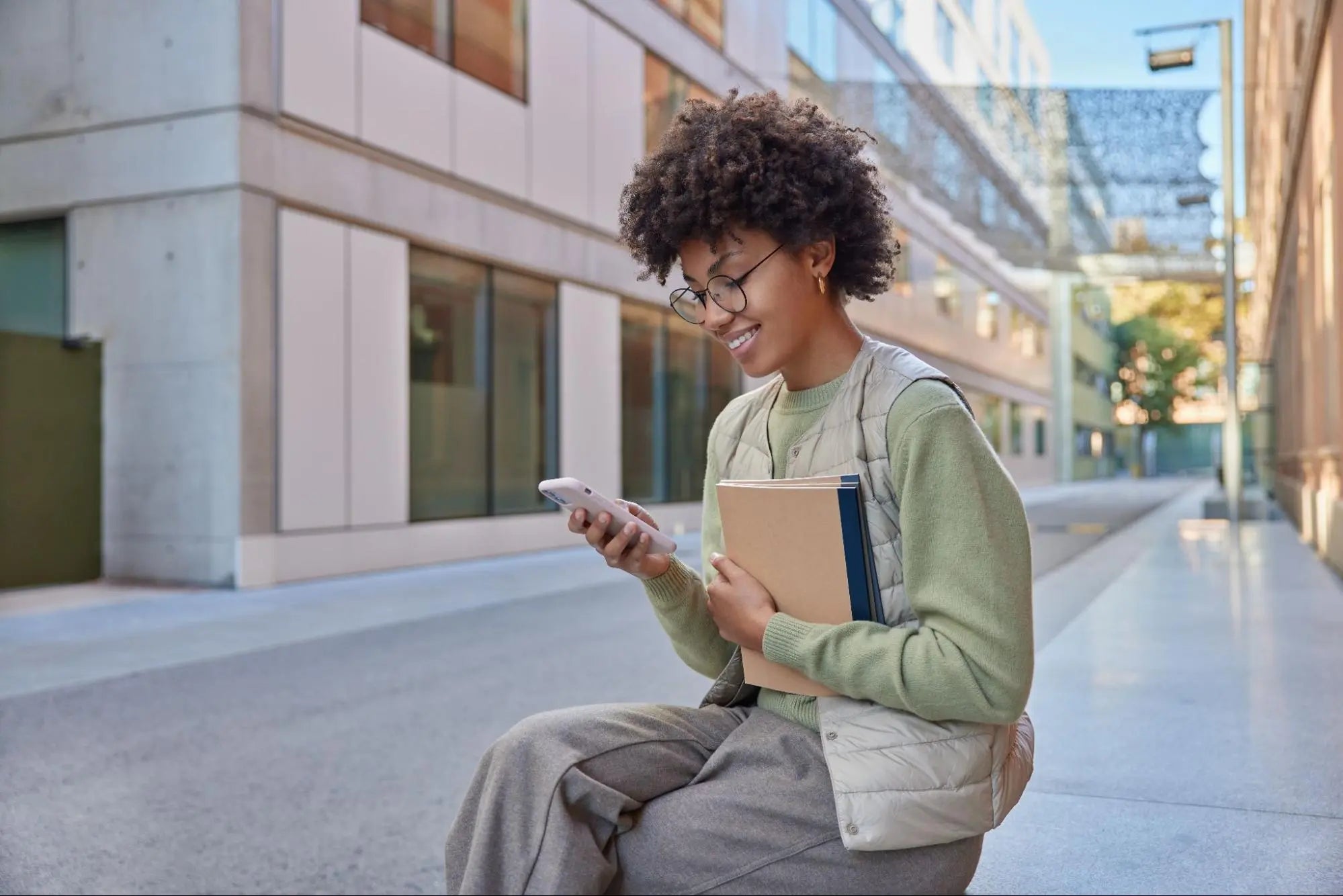 Jovem sorridente com cabelo cacheado e óculos, segurando cadernos e usando um smartphone claro ao ar livre, pesquisando valores e novidades sobre o lançamento de celulares em um ambiente urbano.
