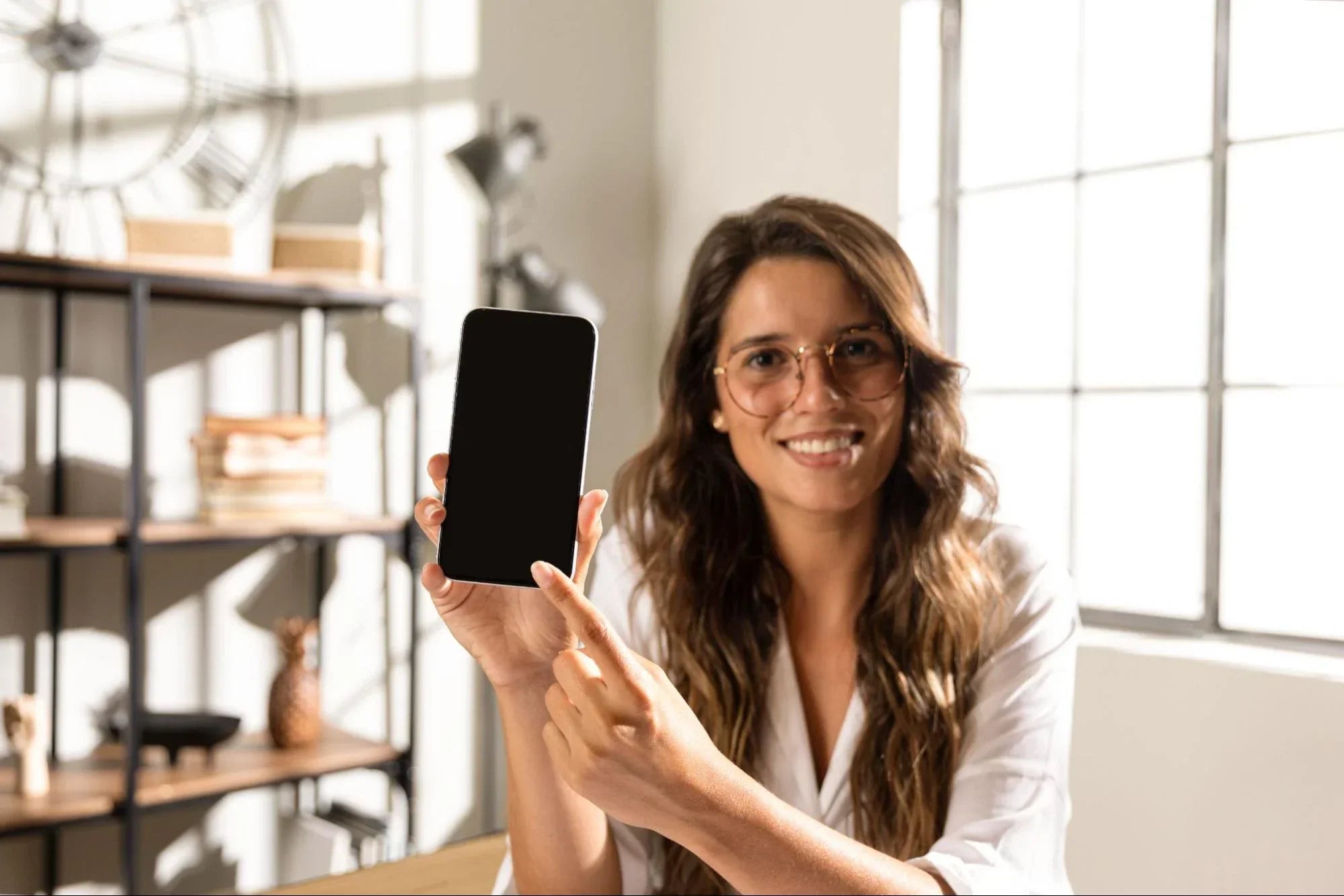 Mulher de óculos sorrindo enquanto segura e aponta para a tela de um smartphone moderno sem bordas, apresentando as novidades de design do aparelho em um ambiente bem iluminado.